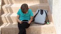 A child sitting on steps using a tablet with a backpack beside them.