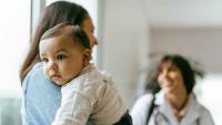 An infant resting on their mother’s shoulder while a clinician looks on.