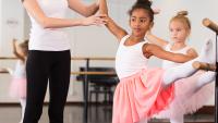 Young girl raises leg on barre during ballet class while instructor helps.