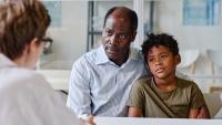 Black father with his little son sitting at table and listening to consultation with doctor at office