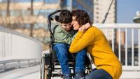 A parent hugs a smiling child sitting in a wheelchair outdoors.