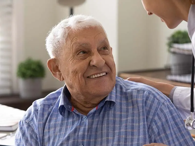 An older man with short white hair in a blue plaid shirt smiling up at a physician in a lab coat with her hand on his shoulder