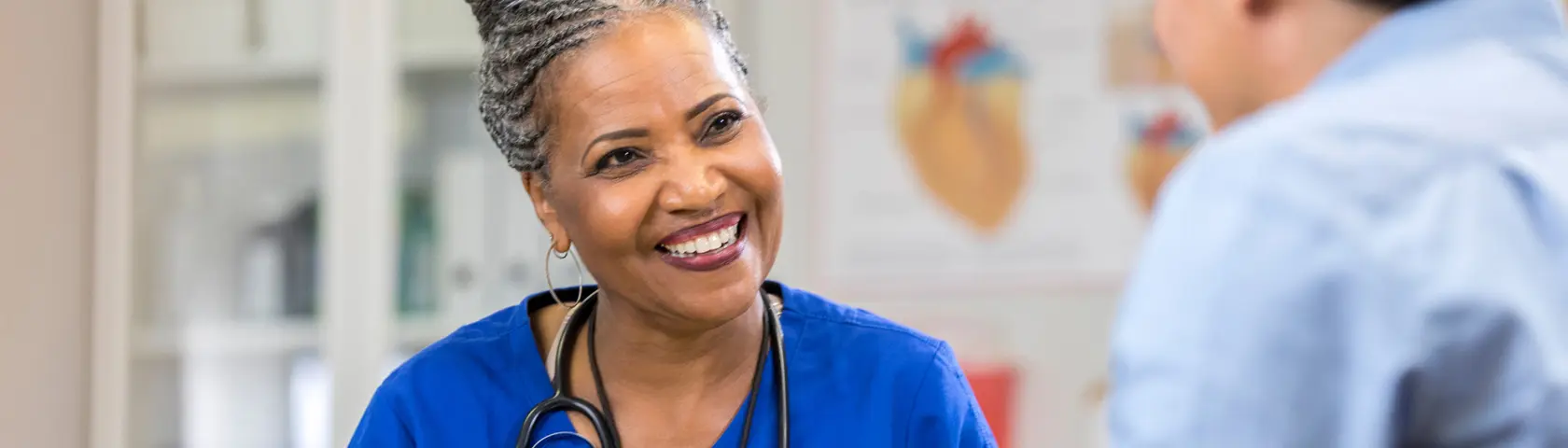 Female black doctor in royal blue scrubs and a stethoscope draped around her neck smiling at a patient in the foreground. Anatomical charts can be seen in the background.