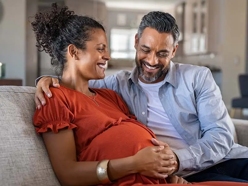 A pregnant woman sitting on a sofa next to a man with his arm draped around her shoulder. Both are smiling clasping each others hands over her belly.