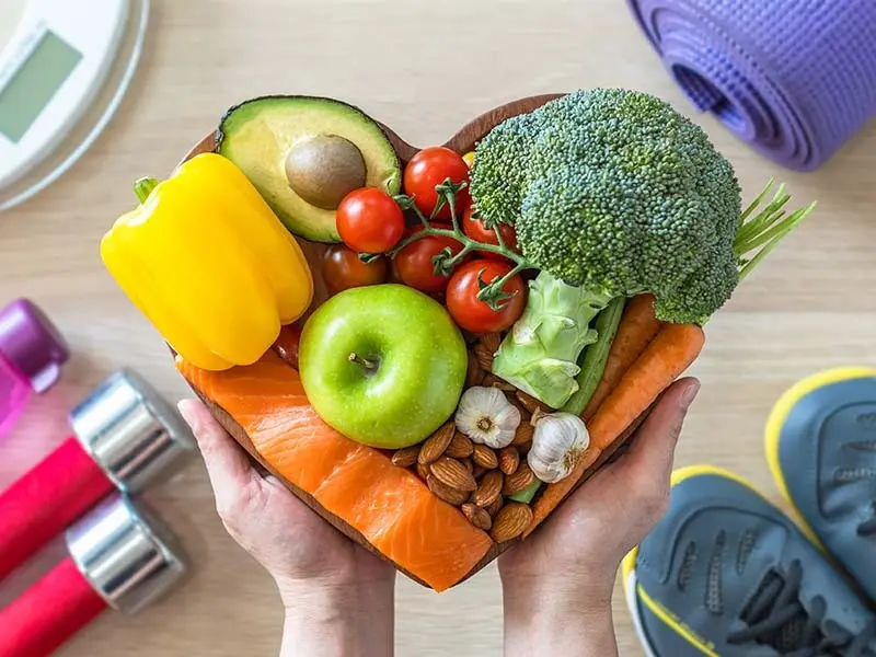 Looking down on hands holding out a heart shaped plate filled with healthy food. In the background a wooden floor with running shoes, a yoga mat. weights and a scale.