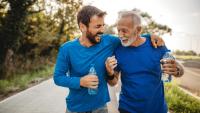 A younger man and an older man, each wearing a blue shirt and holding a bottle of water, laughing while taking a break from jogging