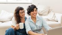 A lesbian couple sit in their living room watching a laptop screen and smiling.