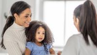 A mother sits at the doctors with her toddler on her lap, as they talk with the doctor.