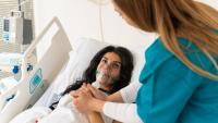 A patient lying in hospital bed with an oxygen mask and holding hands with a nurse