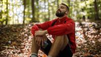 Portrait of young bearded man with bluetooth headphones breathing sitting thinking exercising in woods in park