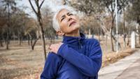 Senior woman holds shoulder and grimaces in pain while walking in park..