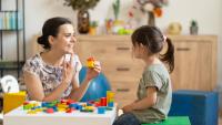 Female autism expert interacting with a young girl during a play-based session, using colorful wooden blocks in a cozy, child-friendly room