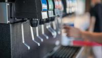 A row of self-service soda dispensers at a beverage station, with a person filling a cup in the background.