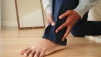 Person kneeling on a wooden floor while holding their lower leg, appearing to experience pain or discomfort near the shin or ankle due to CVI