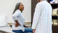A woman sitting on an exam table talks with a clinician while holding her lower back.