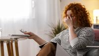 Stressed woman sitting on a couch rubbing her eyes while holding glasses and a notebook at home
