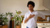 Woman standing in kitchen holding her chest and lower back, appearing to experience chest pain or discomfort at home