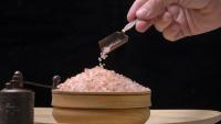 Hand pouring pink Himalayan salt crystals into a wooden bowl, with a salt grinder nearby.