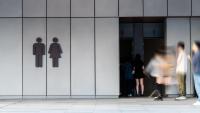 A group of people walking in front of a modern building with a gray facade. The building appears to be a public restroom, as there are two doors on the left side of the image