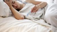 A young woman lying in bed with her head resting on a white pillow. She is wearing a white tank top and appears to be sleeping. A person's hand is visible on the right side of the image, holding a blue thermometer.
