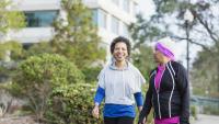 Two African-American women walking hand in hand on a sidewalk. They are both wearing workout clothes and appear to be in a park or garden. The woman on the left is wearing a gray hoodie and black leggings, while the woman in the middle has a pink headband