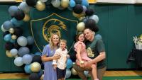 A family of four posing for a photo in front of a large balloon arch. The arch is made up of blue, black, and gold balloons, with a green and gold logo in the center. The family consists of a man, a woman, and two young children.