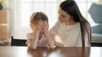 A mother and daughter sitting at a wooden table in a living room. The daughter is covering her face with her hands, while the mother is looking down at her with a concerned expression. The mother is wearing a white t-shirt and has long dark hair.