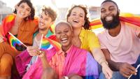A group of five young people, four men and two women, sitting on a bench and smiling at the camera. They are all holding rainbow flags and appear to be at a pride parade or festival. The background shows a cityscape with buildings and a clear blue sky.
