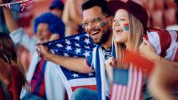 A young couple, a man and a woman, sitting in a stadium and holding American flags. They are both smiling and appear to be happy. The man is wearing glasses and has a beard, while the woman has her face painted in the colors of the American flag.
