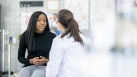Two women in a doctor's office. The woman on the left is sitting on a stool and is wearing a black top and jeans. She has long dark hair and is looking off to the side with a serious expression on her face.