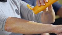 A person's hand holding a yellow spray bottle. The person is wearing a gray t-shirt and appears to be in a park or outdoor setting. The spray bottle is being used to apply sunscreen to the person's arm.