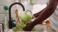 A person's hands washing a large head of cabbage in a kitchen sink. The person is wearing a white t-shirt and is standing in front of a white countertop with a black faucet.