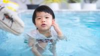 A young child in a swimming pool. The child is in the center of the image, with their body partially submerged in the water. They are wearing a white swimsuit with red and white polka dots. Their hand is on their ear.