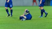 A soccer player lying on the ground with his head in his hands. He is wearing a blue jersey and white soccer cleats. His body is covered in sweat and his arms are stretched out to the sides.