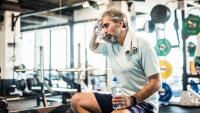 A man sitting on a bench in a gym. He is wearing a light blue t-shirt and black shorts. He has a white towel wrapped around his neck and is holding a water bottle in his left hand.