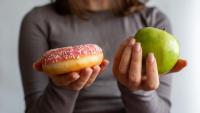 A person holding a donut and a green apple in their hands. The person is wearing a grey long-sleeved shirt and has shoulder-length brown hair. The donut is a light brown color with white sprinkles on top.