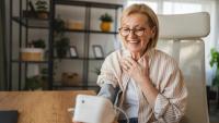 An elderly woman sitting at a desk in an office. She is wearing a striped blouse and glasses and has a smile on her face. The woman is holding a white tablet in her hand and appears to be engaged in a conversation.
