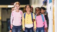 A group of six young children walking in a hallway. They are all wearing backpacks and are smiling at the camera. The girl in the center is wearing a yellow cardigan and blue jeans, while the girl on the left has blonde hair and carries a pink backpac