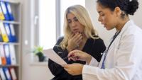 Two women in a medical office. The woman on the left is sitting on a chair and is holding a piece of paper in her hand. She appears to be explaining something to the other woman, who is standing next to her.
