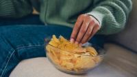 A person sitting on a couch with a bowl of potato chips in front of them. The person is wearing a green sweater and blue jeans. The bowl is transparent and the chips are golden brown in color. The background is blurred, but it appears to be a living room.