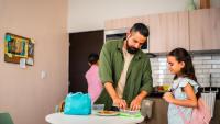 A man and a young girl in a kitchen. The man is standing in front of a round white table with a blue bag on it. He is wearing a green jacket and has a beard. The girl is standing next to him, wearing a pink backpack.