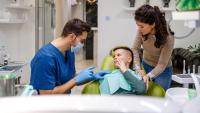 A dentist examining a young boy's teeth in a dental clinic. The dentist is wearing a blue scrub suit and a face mask, and is holding the boy's hand. The boy is sitting in a green dental chair with his mouth open, and his eyes are closed.
