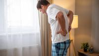 A man standing in a bedroom with his back to the camera. He is wearing a white t-shirt and blue plaid pajama pants. He appears to be in pain or discomfort, with his hands on his lower back and his eyes closed.