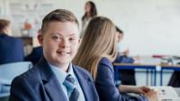 A young boy sitting at a desk in a classroom. He is wearing a blue suit and a red tie. He has short blonde hair and is smiling at the camera. Behind him, there are other students sitting at their desks and a teacher standing in front of a whiteboard