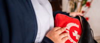 A close-up of a person's hand holding a red first aid kit. The person is wearing a white shirt and a blue jacket. The background is blurred, but it appears to be a room with a vase of red flowers on the wall.