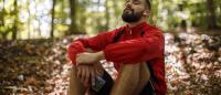 Portrait of young bearded man with bluetooth headphones breathing sitting thinking exercising in woods in park