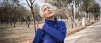 Senior woman holds shoulder and grimaces in pain while walking in park..