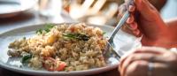 Close-up of a person eating a plate of fried rice with vegetables using a knife and fork, with natural sunlight streaming in and a blurred background of a dining setting.”