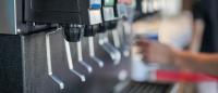 A row of self-service soda dispensers at a beverage station, with a person filling a cup in the background.