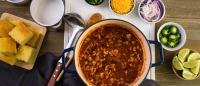 Homemade turkey chili with beans and ground turkey in a pot, served with cornbread, cheese, onions, jalapeños, and lime wedges on a wooden table